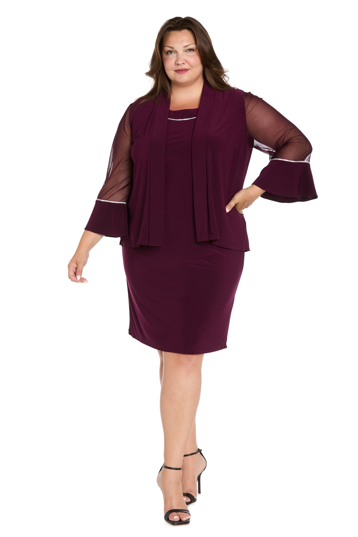 A woman models the R&M Richards 3747W Short Plus-Size Jacket Party Dress, a knee-length deep purple design with sheer long sleeves and a matching overlay, styled with black heeled sandals against a white background.